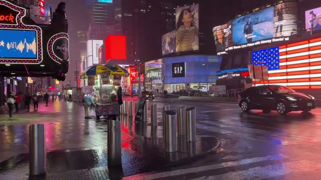 Times Square at night in the rain, with neon billboards and an American flag on a screen